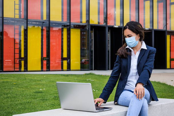 Молодая женщина работает за компьютером в парке с маской во время пандемии - Фото, изображение
