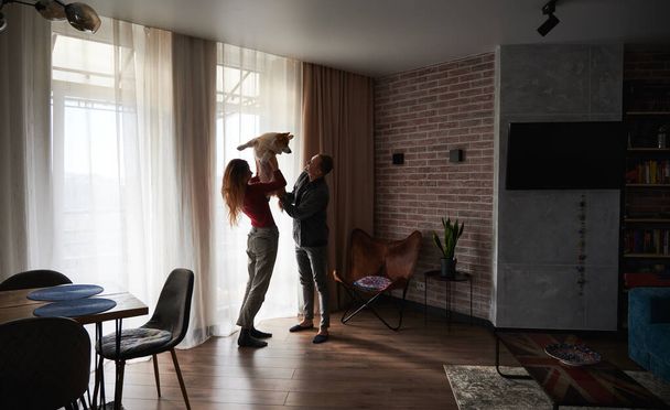 Side view of young woman and man standing and playing with lovely dog at home in kitchen with beautiful interior. Concept of free time together with lovely pet at home. - Foto, afbeelding