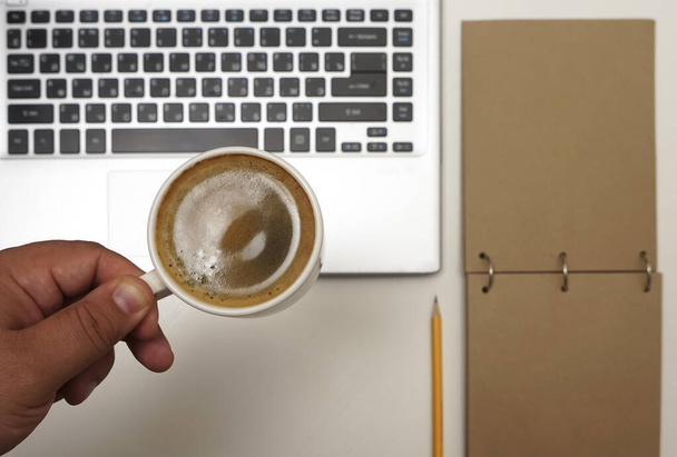  Vista superior da mão masculina com xícara de café acima do laptop. caderno marrom vazio com lápis amarelo.  - Foto, Imagem