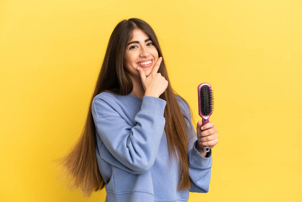 Young caucasian woman holding hairbrush isolated on blue background happy and smiling - Foto, afbeelding
