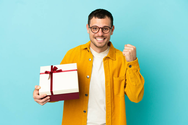 İzole edilmiş mavi arka planda bir hediye taşıyan Brezilyalı adam galibiyeti kutluyor. - Fotoğraf, Görsel