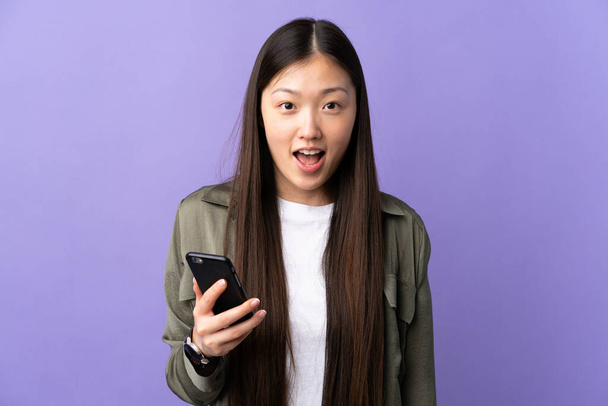 Junge Chinesin benutzt Mobiltelefon über isoliertem lila Hintergrund mit Überraschung und schockiertem Gesichtsausdruck - Foto, Bild