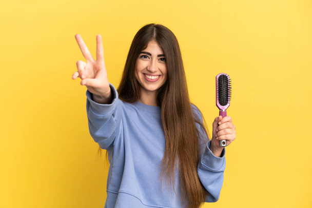 Young caucasian woman holding hairbrush isolated on blue background smiling and showing victory sign - Photo, Image