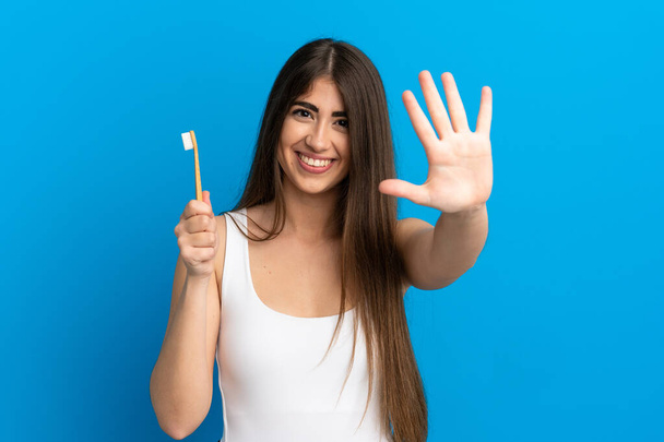 Молодая белая женщина чистила зубы на синем фоне, считая пять пальцев. - Фото, изображение
