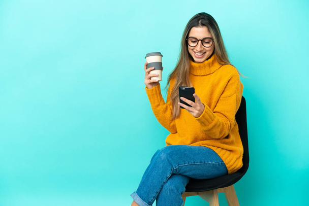 Junge kaukasische Frau sitzt isoliert auf einem Stuhl auf blauem Hintergrund und hält Kaffee zum Mitnehmen und ein Handy in der Hand - Foto, Bild