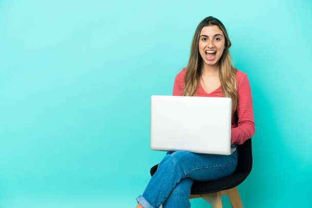Молода біла жінка сидить на стільці зі своїм комп'ютером ізольовано на синьому фоні з несподіваним виразом обличчя
 - Фото, зображення