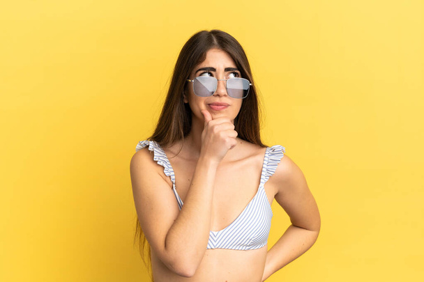 Young caucasian woman in swimsuit in summer holidays isolated on yellow background having doubts and thinking - Zdjęcie, obraz