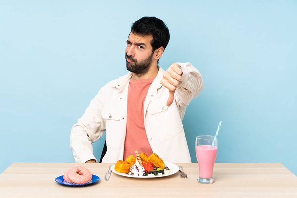 Egy férfi az asztalnál reggelizik, gofrit eszik, és turmixol, negatív arckifejezéssel mutatja a hüvelykujját. - Fotó, kép