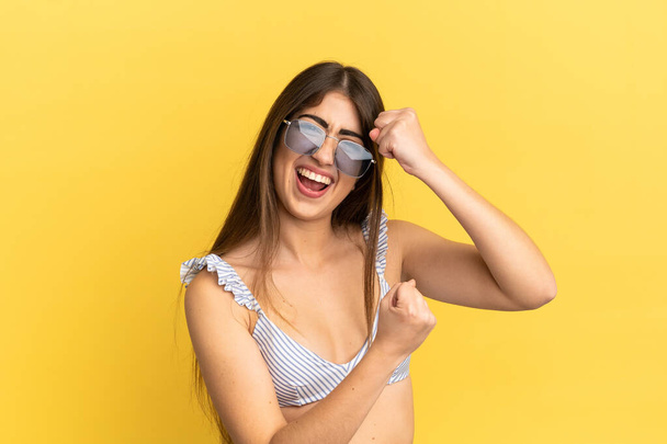 Young caucasian woman in swimsuit in summer holidays isolated on yellow background celebrating a victory - Photo, Image