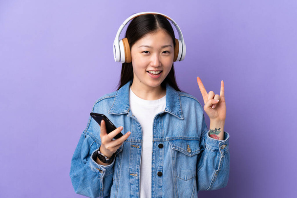 Junges chinesisches Mädchen vor isoliertem lila Hintergrund hört Musik mit einem Handy, das Rock macht - Foto, Bild
