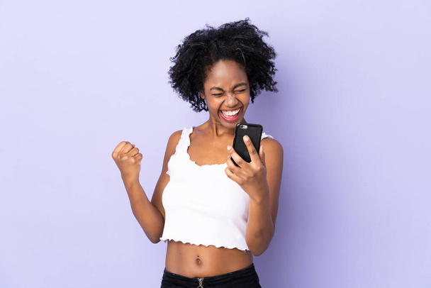 Молодая афроамериканка изолирована на фиолетовом фоне с телефоном в победной позиции - Фото, изображение