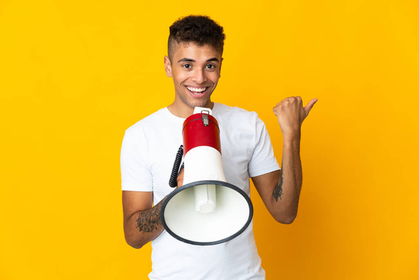 Jeune homme brésilien isolé sur fond jaune criant à travers un mégaphone et le côté pointant - Photo, image