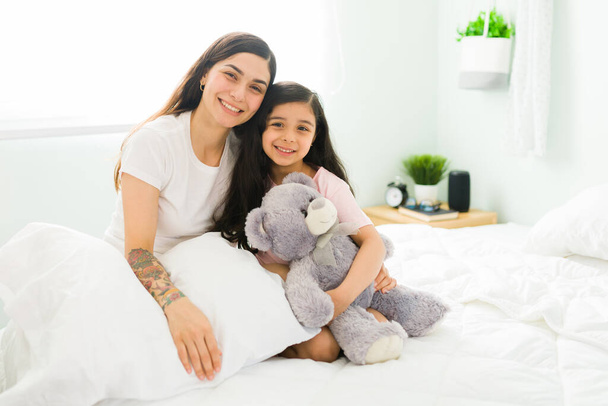 Portrait of a beautiful young mother and her cute daughter with a teddy bear hugging while sitting together in a comfy white bed - Fotoğraf, Görsel