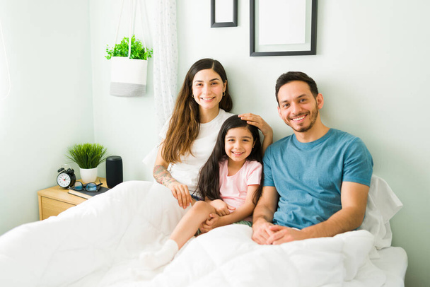 Porträt einer jungen glücklichen Familie, die am Wochenendmorgen zusammen im Bett liegt. Hispanische Mutter, Vater und Tochter fühlen sich glücklich und entspannt - Foto, Bild