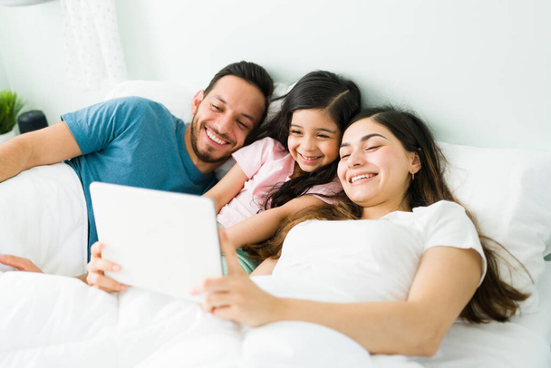 Feliz joven mamá y papá riendo con su pequeña hija en el dormitorio mientras ve videos de comedia o películas en una tableta  - Foto, Imagen