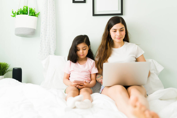 Attractive mother working and typing on a laptop while sitting with pajamas in bed. Little daughter resting next to her mom and using a smartphone - Photo, Image