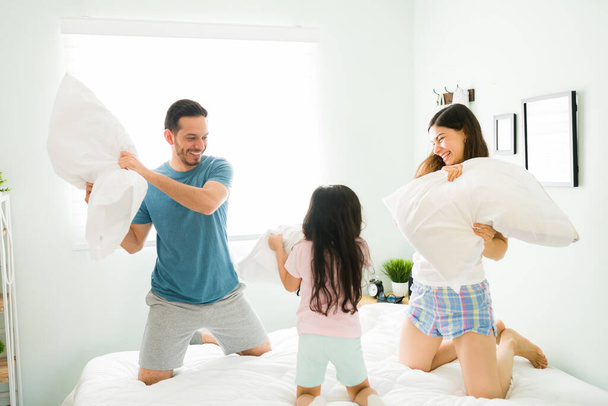 Uma menina feliz a começar uma luta de almofadas com os pais na cama. Mãe e pai brincando enquanto se divertindo com sua filha brincalhona - Foto, Imagem