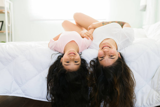 Playful young latin mom and little girl with beautiful long hair lying upside down in bed while smiling - Фото, зображення