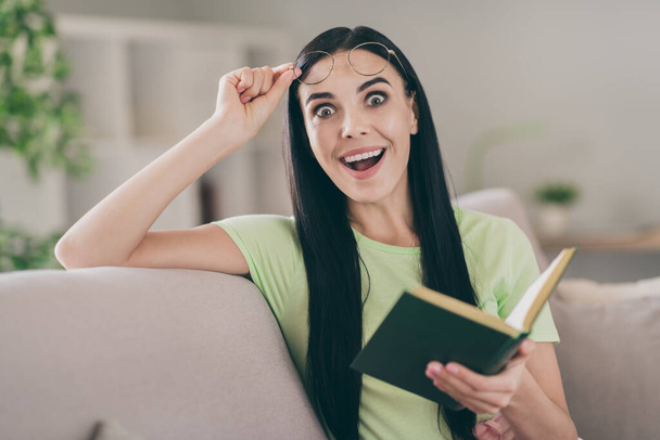 Photo of funny excited girl sit sofa hold book take off specs open mouth wear eyeglasses green t-shirt indoors - Valokuva, kuva