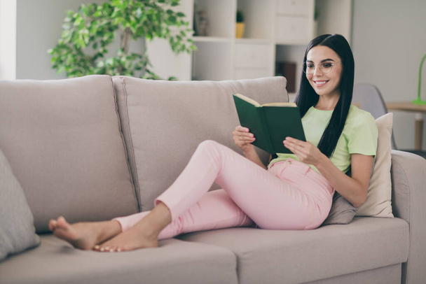 Photo of cheerful clam young lady lay sofa pillows hold book read barefoot wear glasses green t-shirt pink trousers indoors - Fotó, kép