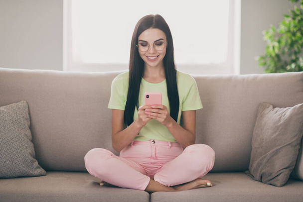 Photo of cheerful young girl sit sofa crossed legs hold telephone look screen toothy smile wear specs green t-shirt pink pants indoors - Fotoğraf, Görsel