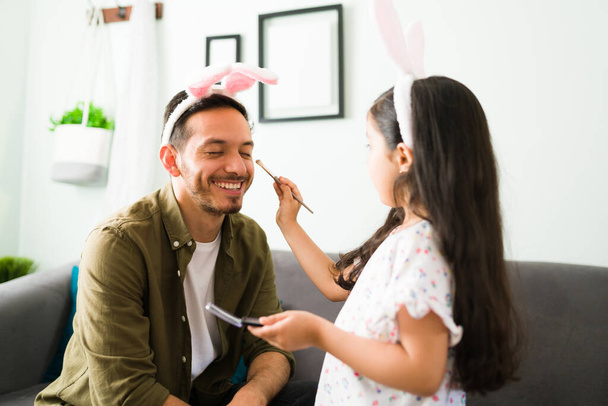 Cheerful dad with bunny ears playing with her little daughter. Beautiful girl putting makeup and eyeshadow to her father - Zdjęcie, obraz