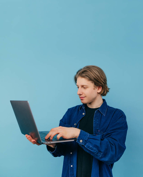 Young male freelancer in roe deer clothes stands with a laptop in his hands on a blue background and works with a smile on his face. Vertical. - Zdjęcie, obraz