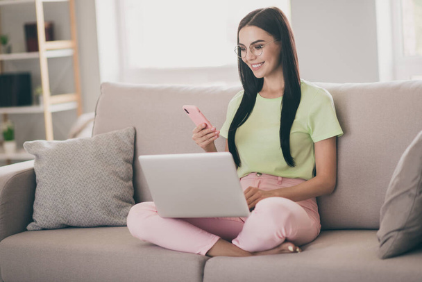 Photo of cheerful girl sit sofa crossed legs hold phone netbook wear spectacles green t-shirt pink trousers indoors - Fotó, kép
