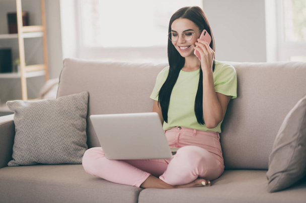 Photo of charming girl sit couch hold phone conversation laptop look screen wear glasses green t-shirt pink pants indoors - Foto, afbeelding