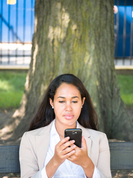 Mieszana rasa afro-amerykańska bizneswoman w brązowej lub beżowej marynarce siedząca pod drzewem cieni na ławce w parku patrzy na swój telefon w słoneczny letni dzień. - Zdjęcie, obraz