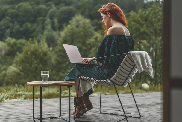 Πίσω όψη της επιτυχημένης γυναίκας που εργάζονται με φορητό υπολογιστή και κάθεται σε εξωτερικό χώρο σε μια όμορφη βεράντα στο δάσος - Φωτογραφία, εικόνα