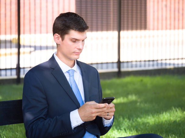 Valkoinen valkoihoinen liikemies tummansinisessä puvussa ja solmio tekstejä kännykkään istuessaan puistossa ruoho, tiili ja aita taustalla.. - Valokuva, kuva