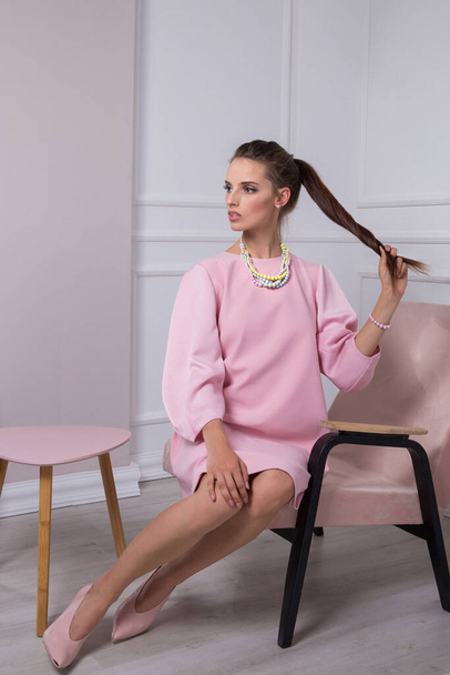 Beautiful brunette girl in a pink dress sits on a chair pulling a braid with a raised hand. - Fotografie, Obrázek