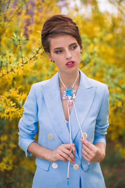 Young brunette girl model in a blue jacket in the park. Woman with beautiful crystal necklace. Natural background. - Foto, afbeelding