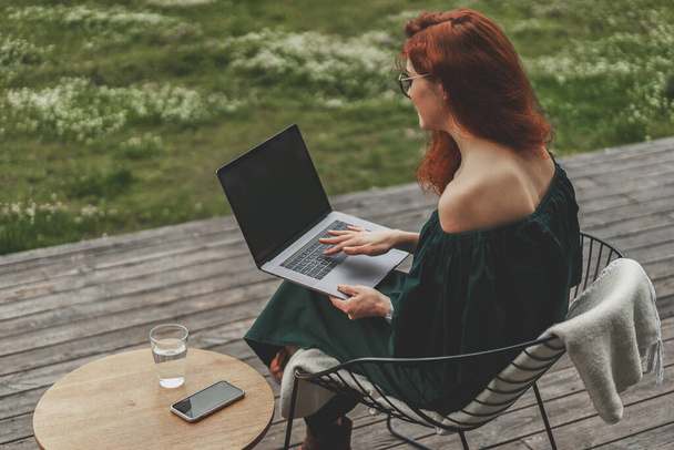 Вид сзади на бизнес-женщину, работающую с ноутбуком и сидящую на открытой террасе в лесу - Фото, изображение
