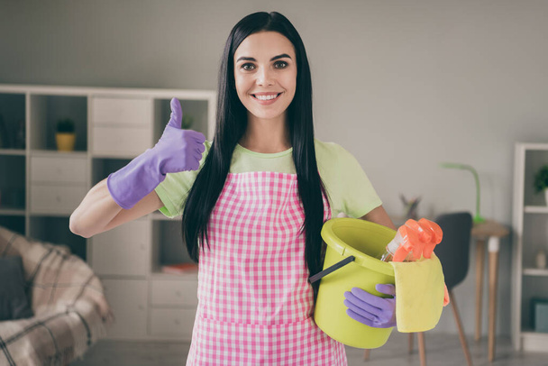 Portrait of attractive content cheerful long-haired maid holding in hands bucket with supplies showing thumbup at light interior living-room - Photo, image