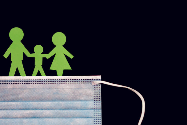 Schutzmaske vor Infektionen und Männer aus grünem Papier in Form einer Familie auf schwarzem Hintergrund. Die Familie trägt eine Maske gegen Coronavirus und andere Infektionen durch die Luft. Freiraum für Text - Foto, Bild