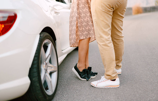 Chica en un vestido colorido se encuentra de puntillas cerca de hombre en pantalones amarillos contra el telón de fondo de un hermoso coche blanco. Primer plano de las piernas - Foto, Imagen