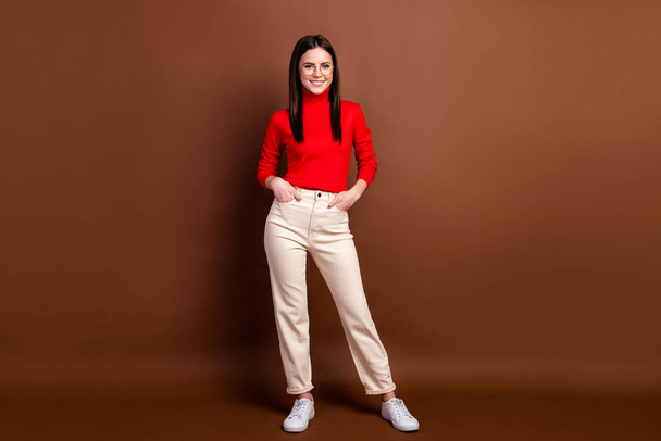 Photo of pretty charming young woman dressed red sweater standing arms pockets smiling isolated brown color background - Fotó, kép