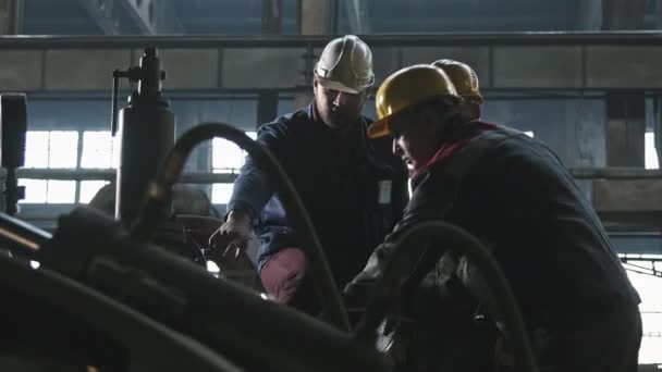 Średnie ujęcie trzech pracowników ciągników męskich w kombinezonach i kaskach stojących przy sprzęcie maszynowym w odlewni omawiających pracę - Materiał filmowy, wideo
