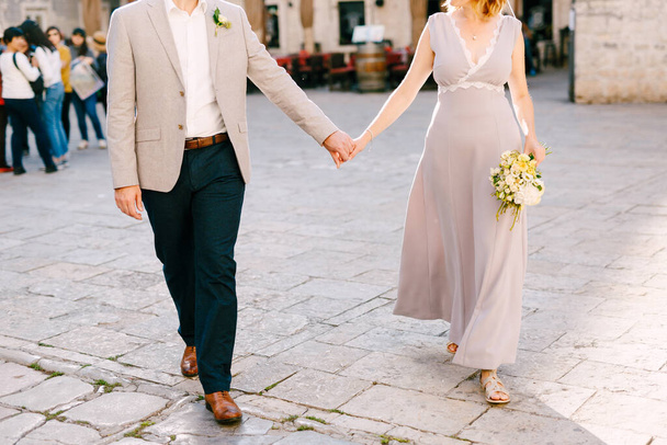 Νύφη και γαμπρός κρατιούνται χέρι-χέρι στις πλακόστρωτες πέτρες της πλατείας - Φωτογραφία, εικόνα