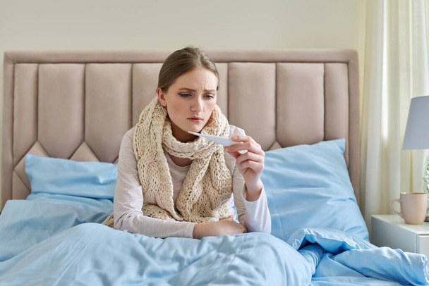 Sick young woman with thermometer in her hands, sitting at home in bed - Фото, изображение