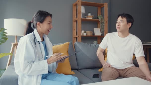 Mladá Asie žena profesionální lékař pomocí digitální tablet sdílení dobré zprávy o zdravotním testu s šťastný mužský pacient sedět na gauči v domě. Zdravotní pojištění, Návštěva pacienta doma koncepce. - Záběry, video