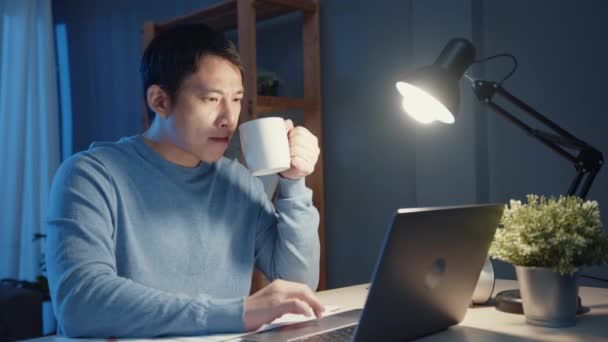 Азіатський бізнесмен відпочиває з чашкою кави і перевіряє роботу на ноутбуці комп'ютера для порядку денного у вітальні в понаднормовий час, роботу з домашньої пандемії корони.. - Кадри, відео