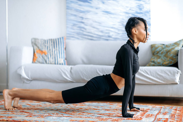 Attraktive aktive schlanke Afroamerikanerin in schwarzer Sportbekleidung, macht Stretching zu Hause auf dem Boden, betreibt engagierte Fitness, führt einen gesunden Lebensstil, kümmert sich um die Gesundheit - Foto, Bild