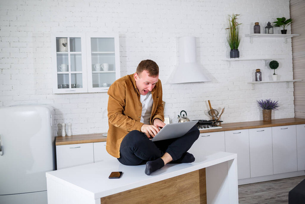 An attractive young man in casual clothes sitting in the kitchen using a laptop computer. Work from home, remote workflow. - Foto, Bild