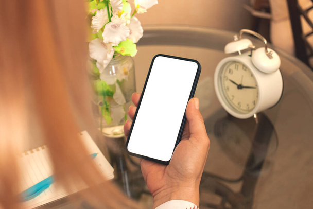 Mujer foto burla celebrar teléfono inteligente moderno con pantalla blanca en blanco en la planta de fondo y reloj despertador - Foto, Imagen