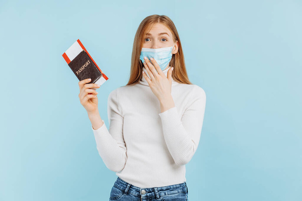 Shocked frightened young woman, in medical mask, holding passport with tickets, on blue background - Фото, изображение
