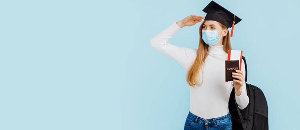 Χαρούμενη νεαρή φοιτήτρια με ιατρική μάσκα, φοιτήτρια με καπέλο αποφοίτησης με διαβατήριο και εισιτήρια σε μπλε φόντο, σπουδές, εκπαίδευση, πανεπιστήμιο, εισαγωγή - Φωτογραφία, εικόνα
