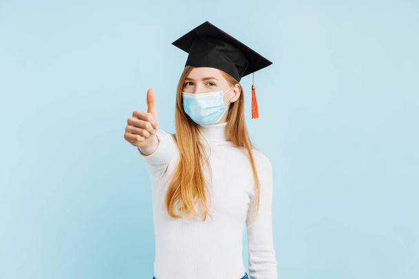 Happy female student graduate in medical mask showing thumb up on blue background - Zdjęcie, obraz